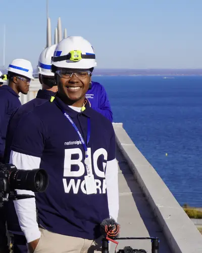 Myles Braxton on location in a hard hat, camera rig in hand, waterfront behind him